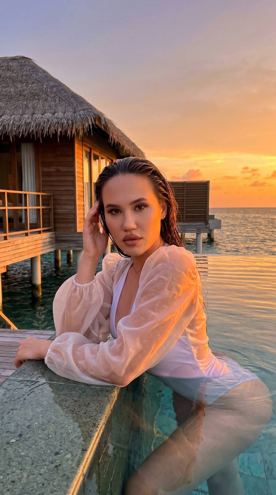 A girl in a white swimsuit and organza cape poses in an infinity pool at sunset, looking confidently at the camera.