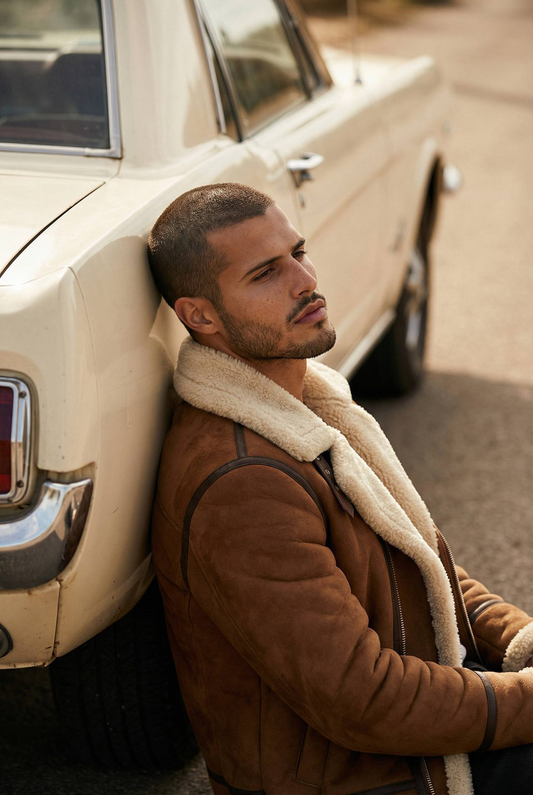 A young man in a brown suede shearling jacket leans against a vintage cream car, gazing thoughtfully off-camera.