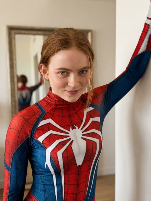 A woman in a red and blue superhero costume posed indoors against a white wall, with light-colored wooden flooring and a mirror reflecting her back.
