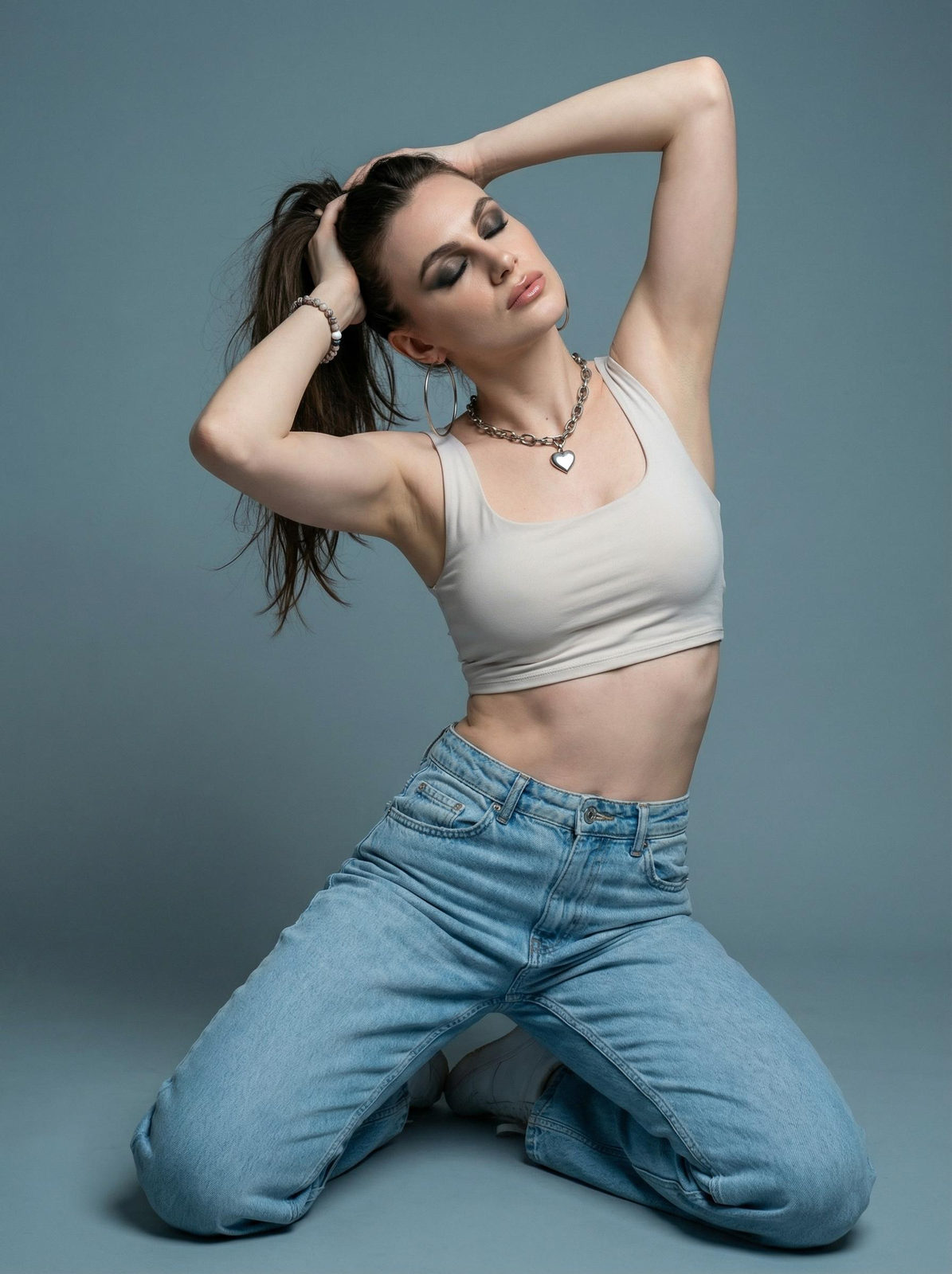 Portrait of a woman kneeling in a minimalist studio, wearing a crop top and jeans, with smoky eye makeup and a high ponytail.