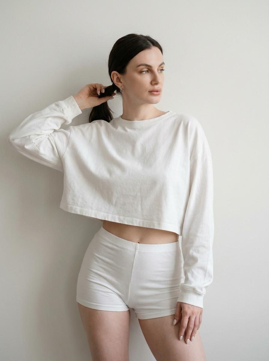 A woman showcasing a minimalist pose against a light wall, featuring natural makeup, a ponytail, and soft lighting.