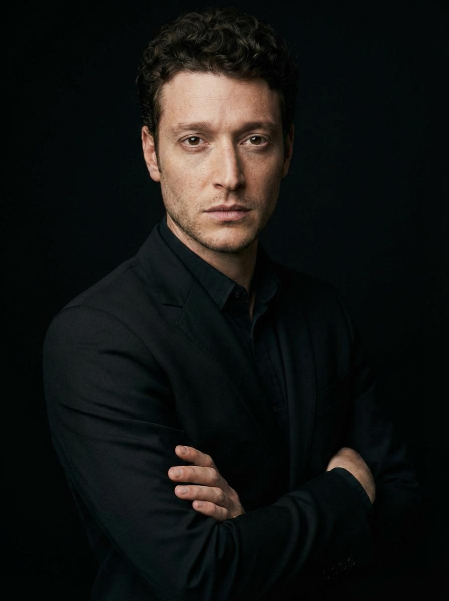 A man in a fitted black suit standing with crossed arms, shot in a 3/4 angle against a solid black background.