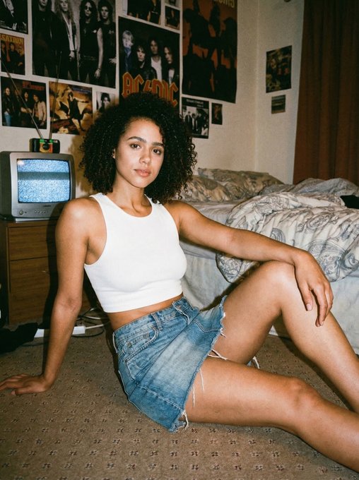 A woman in a white ribbed tank top and distressed denim mini skirt sitting on a carpeted floor in a vintage band poster bedroom.