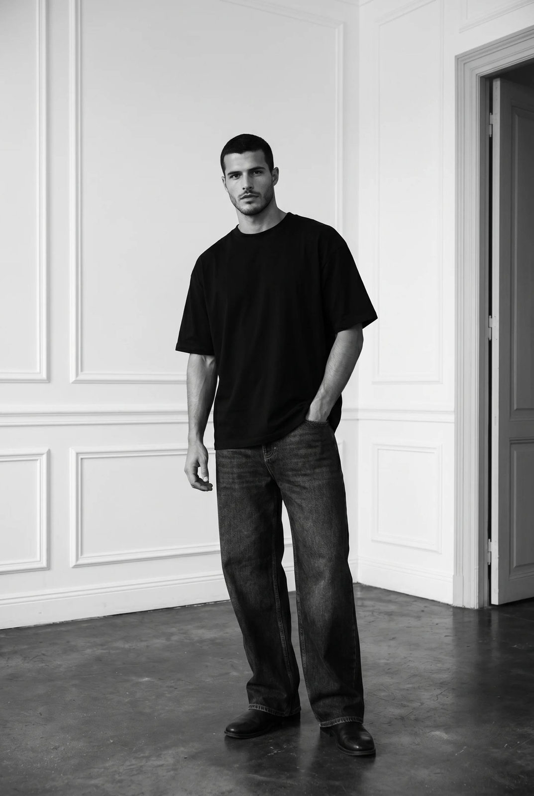 A man in a minimalist interior, casually standing with one hand in pocket, wearing an oversized black t-shirt and dark denim, under soft cinematic lighting.