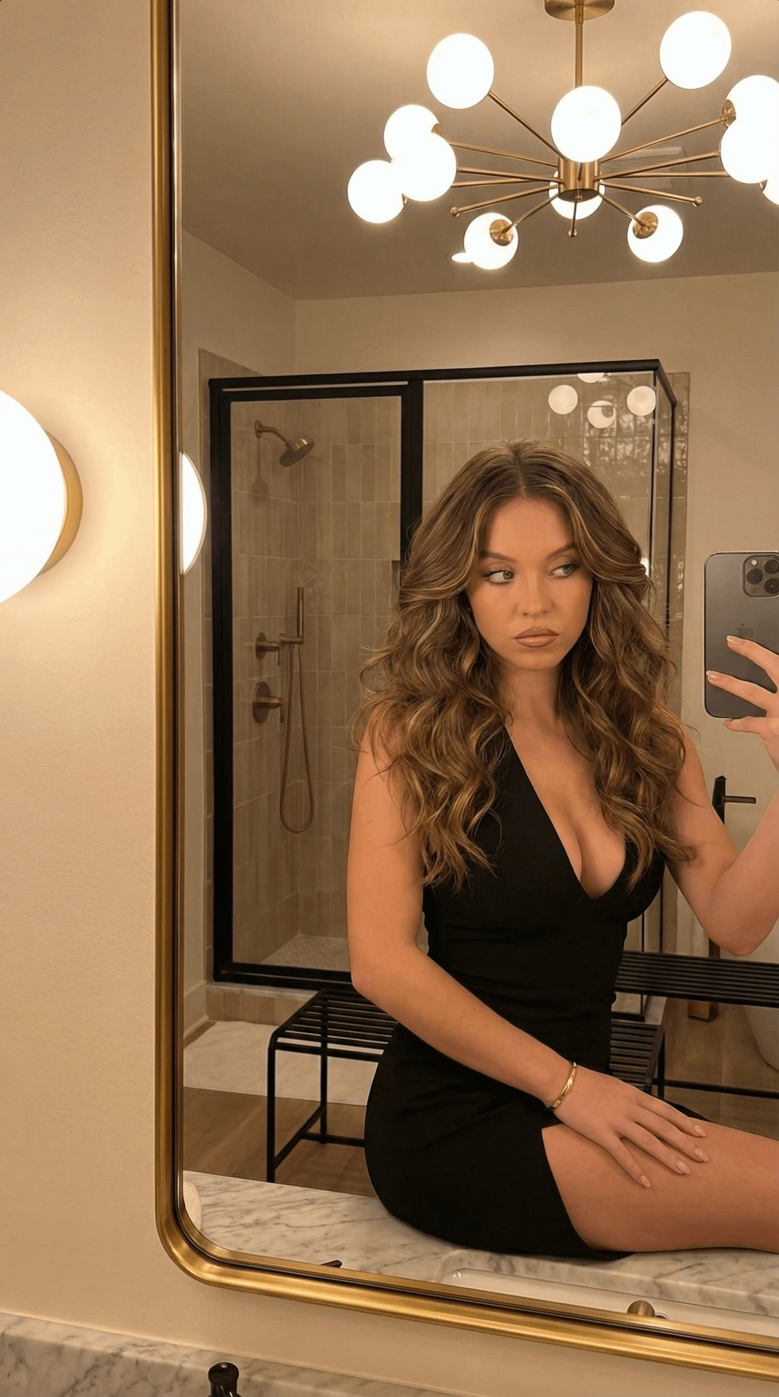 A person taking a bathroom selfie while wearing a black mini dress, seated on a marble countertop with a gold chandelier in the background.