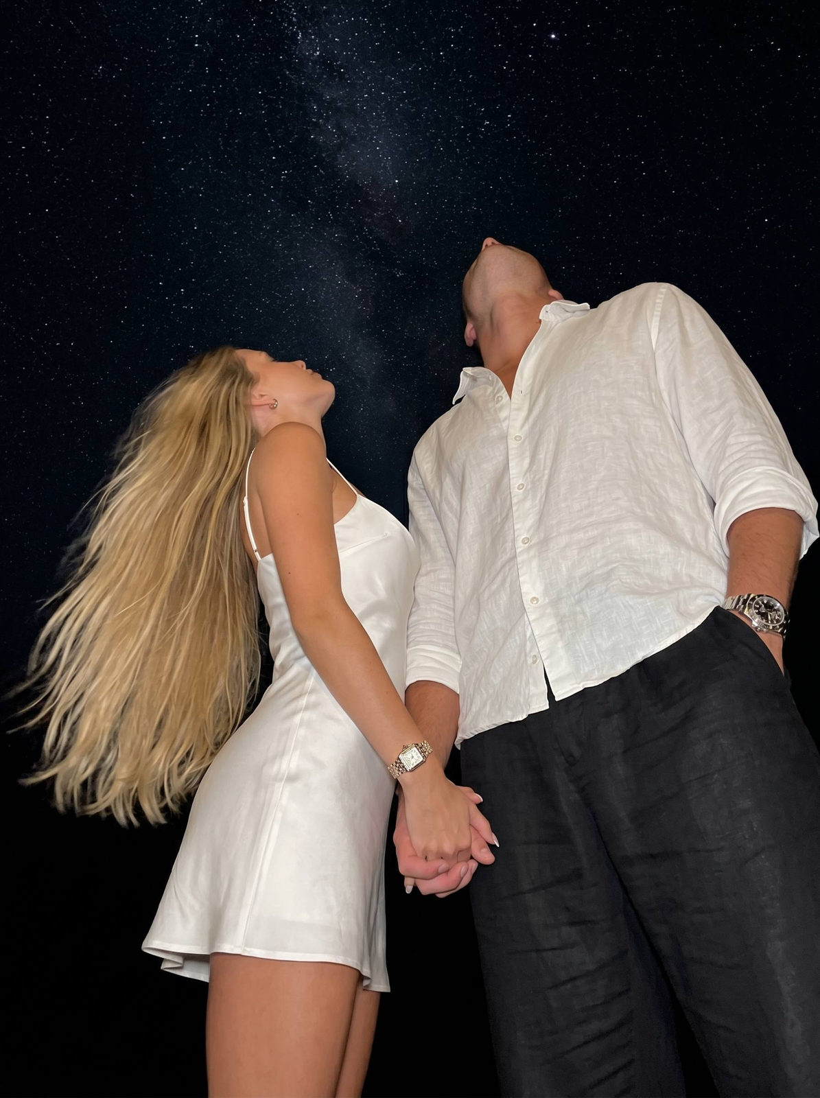 A woman in a white silk slip dress looking up at the Milky Way with a man beside her, both in low-angle view.