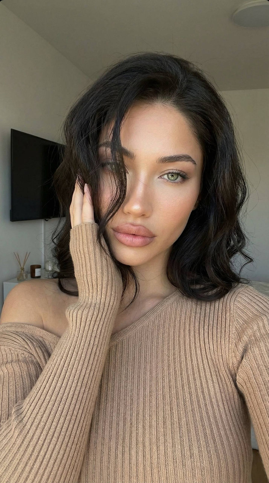 A close-up selfie of a woman in a warm-toned fitted knit top, relaxed pose with soft lighting in a minimalistic room.