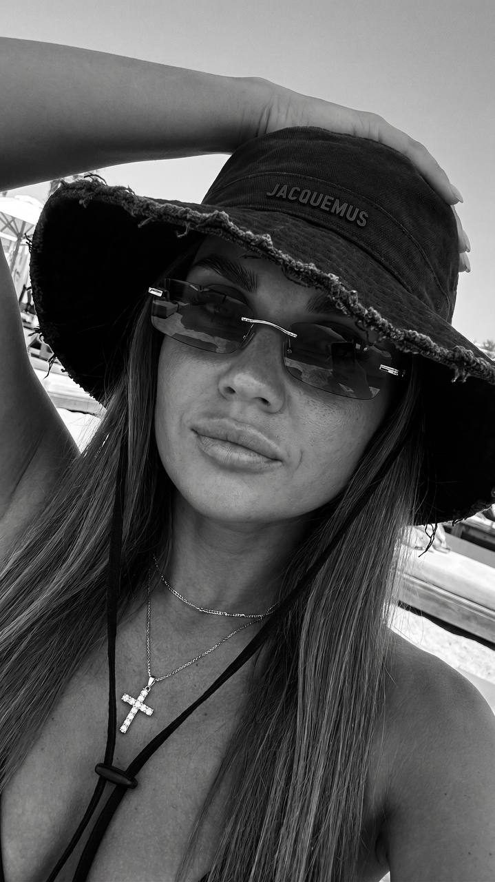 Close-up outdoor selfie of a woman with a deep sun-kissed tan, wearing a crystal cross pendant and oversized black bucket hat in a beach club.