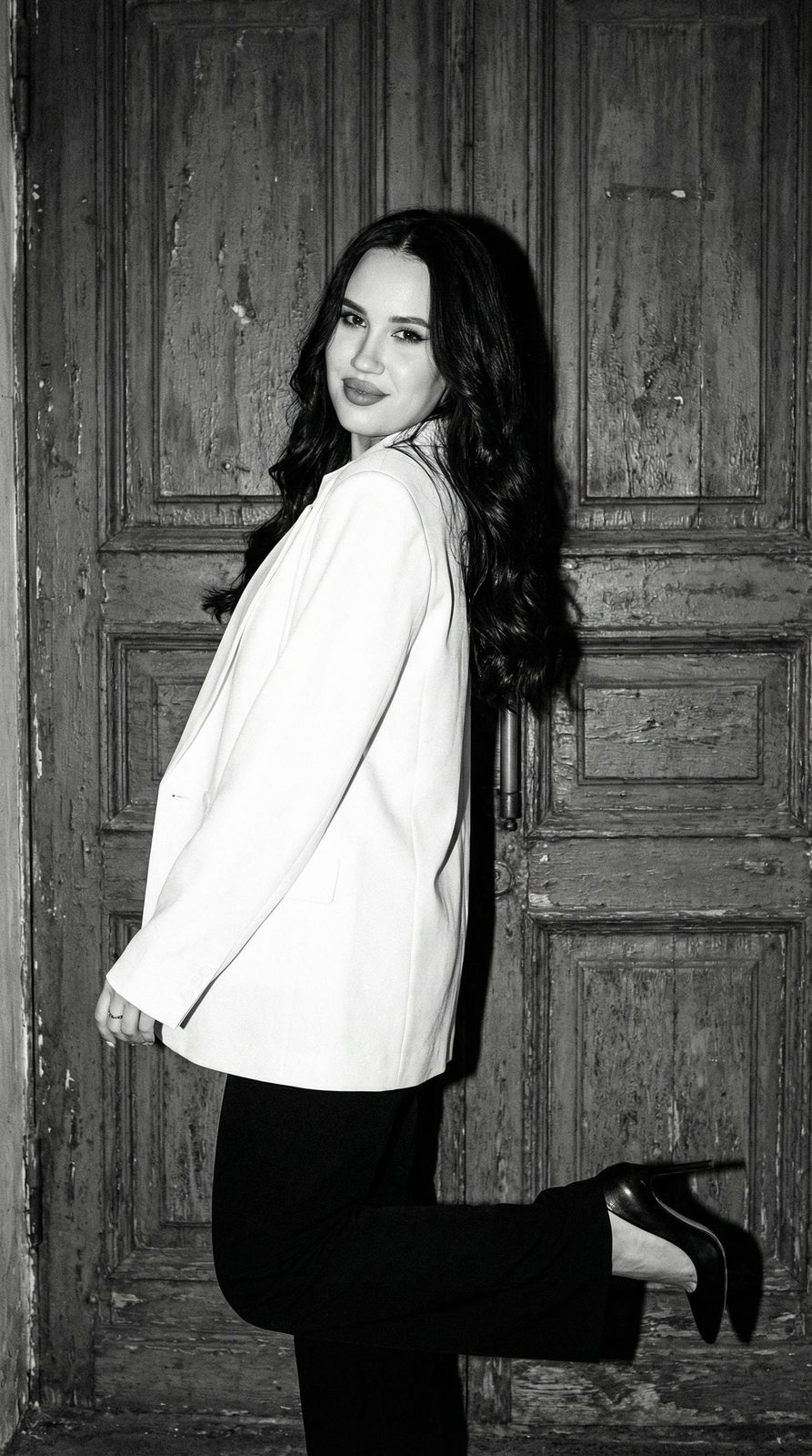 A fashionable girl in an oversized white jacket poses playfully against a textured old door.