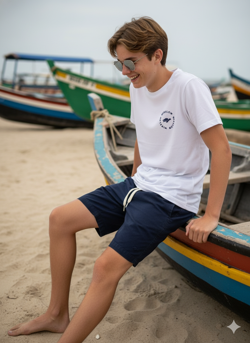 Adolescente de 15 anos sorrindo enquanto sentado na borda de um barco colorido na praia, usando óculos escuros e roupa azul.