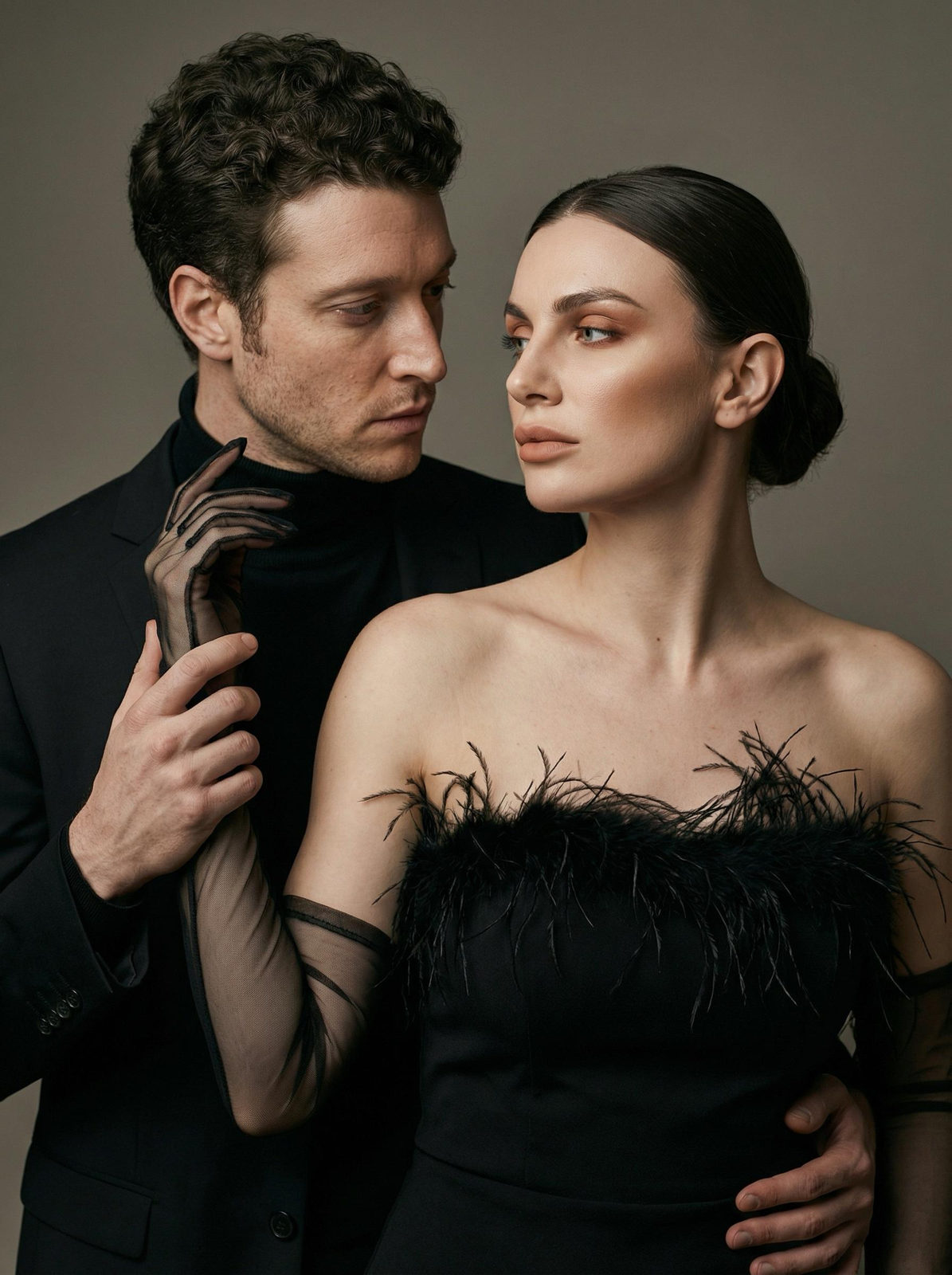 A photorealistic studio portrait of a couple, framed against a soft gray-beige wall. The woman in a strapless dress with delicate feather details, holding hands with the man in a sleek suit.