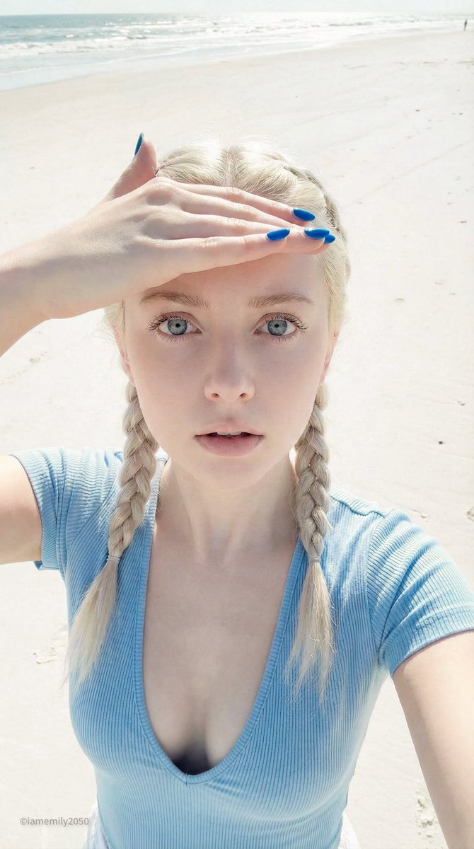 High-angle selfie of a young woman on a sandy beach, shielding eyes from bright light, showcasing porcelain skin and vibrant blue nails.