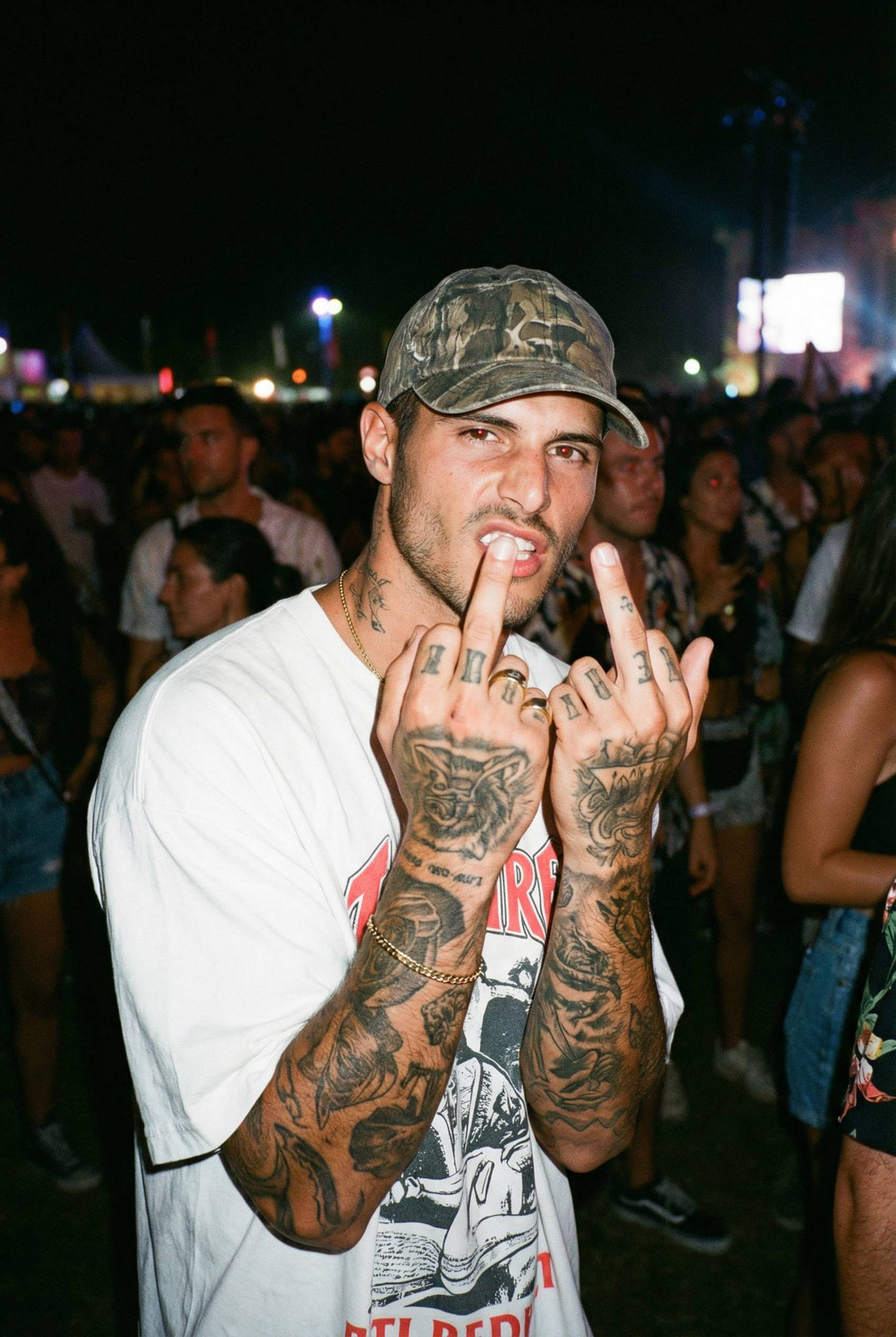 A tattooed man at a nighttime party, confidently raising his hands while wearing a camouflage cap and graphic T-shirt with harsh flash lighting.