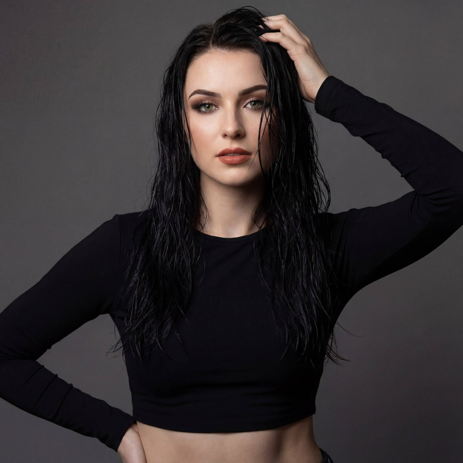 A confident woman with long dark wet hair in a studio portrait, wearing a cropped long-sleeved top, set against a neutral dark gray background.