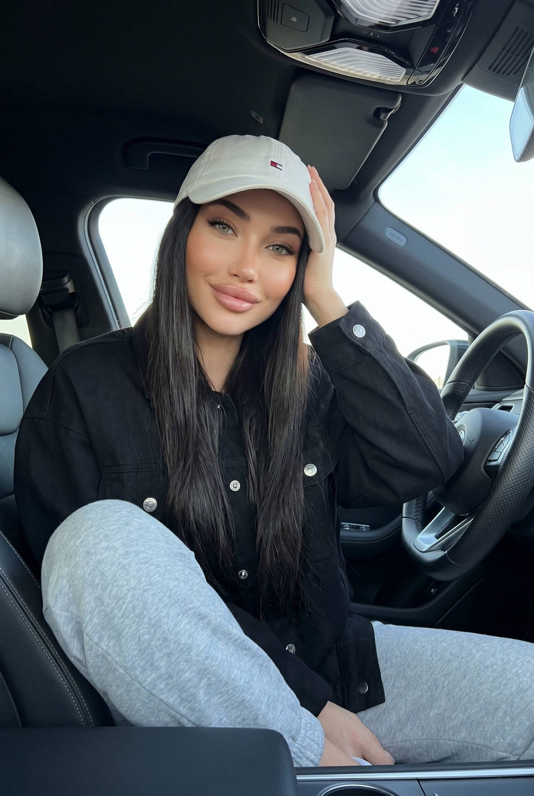 A woman in a white cap and black jacket relaxing in a modern car, casually holding her head with one hand.