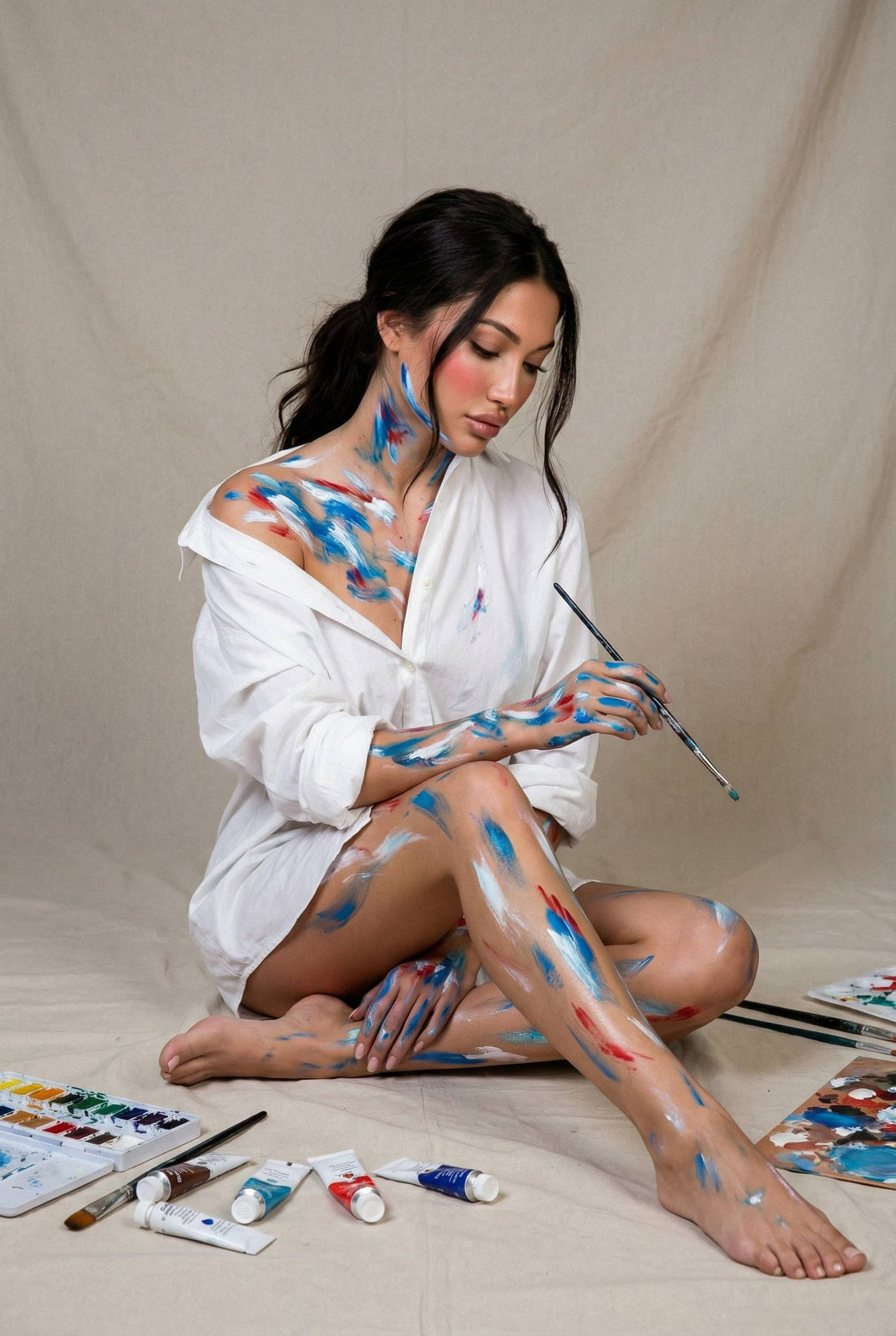 A woman seated on a neutral fabric floor, surrounded by paint tubes and palettes, wearing an oversized white shirt, with abstract paint strokes on her skin.