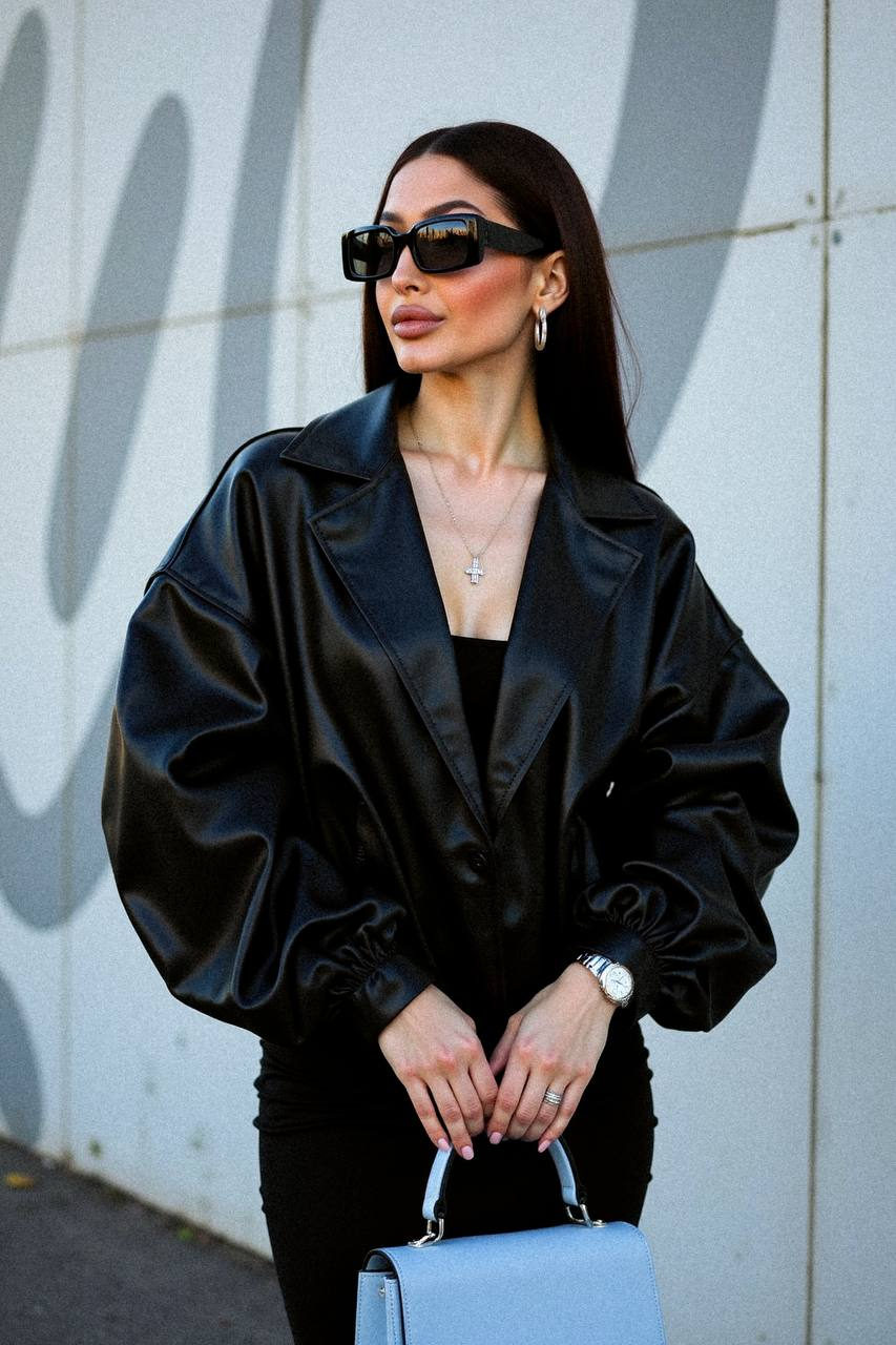 A stylish woman in an oversized leather jacket poses confidently against a modern architectural wall.