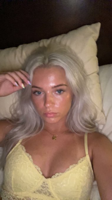A woman with long platinum hair in a pale yellow lace bralette lying on a bed, one hand on forehead, looking towards the camera.
