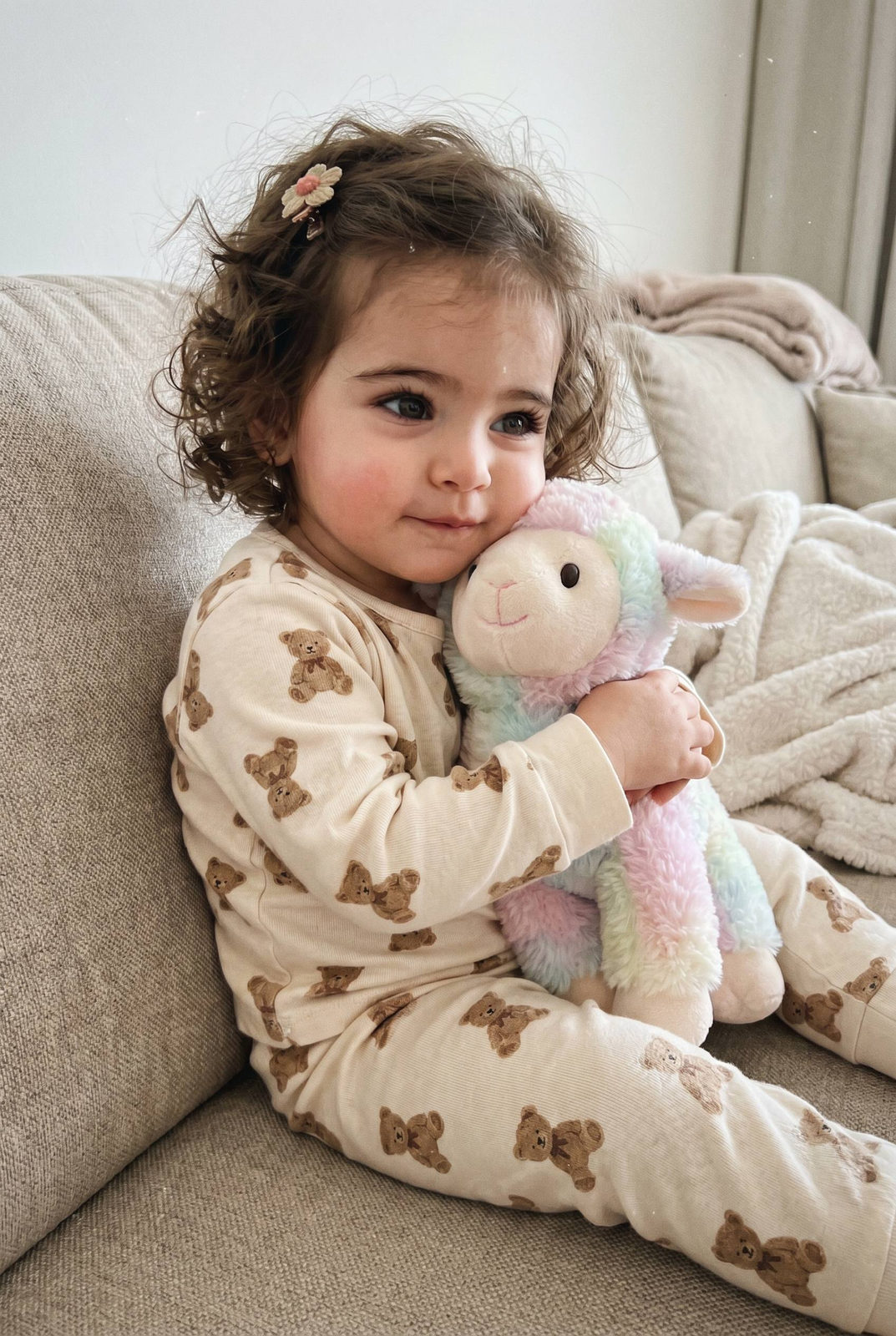Toddler girl in cream pajamas hugging a plush lamb on a couch, with soft hair and a playful expression.