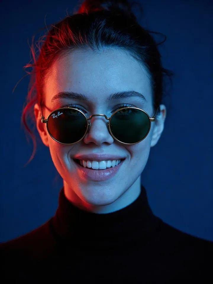 A cinematic portrait of a woman wearing vintage round sunglasses, set against a dark navy background with dramatic lighting.