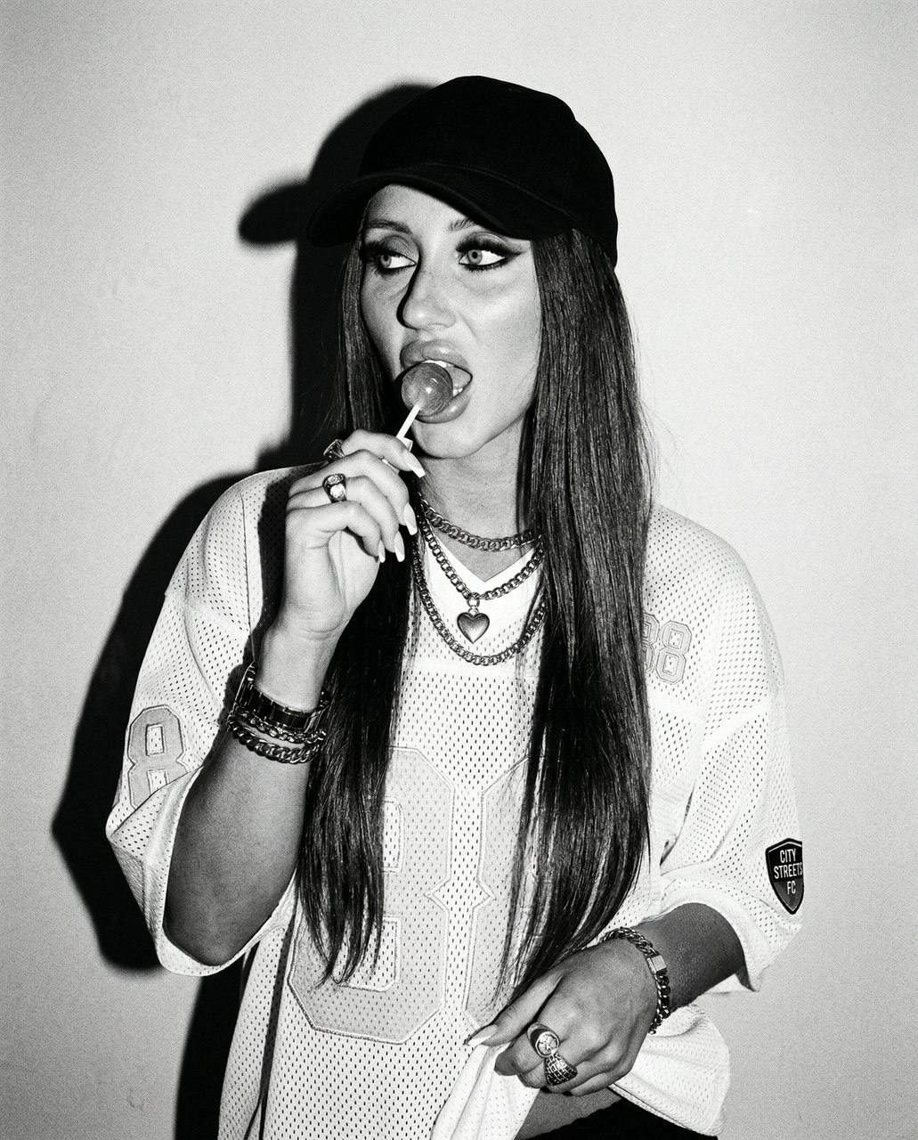Girl in street fashion holding a lollipop, wearing a white mesh jersey and dark baseball cap, with expressive makeup and chains.