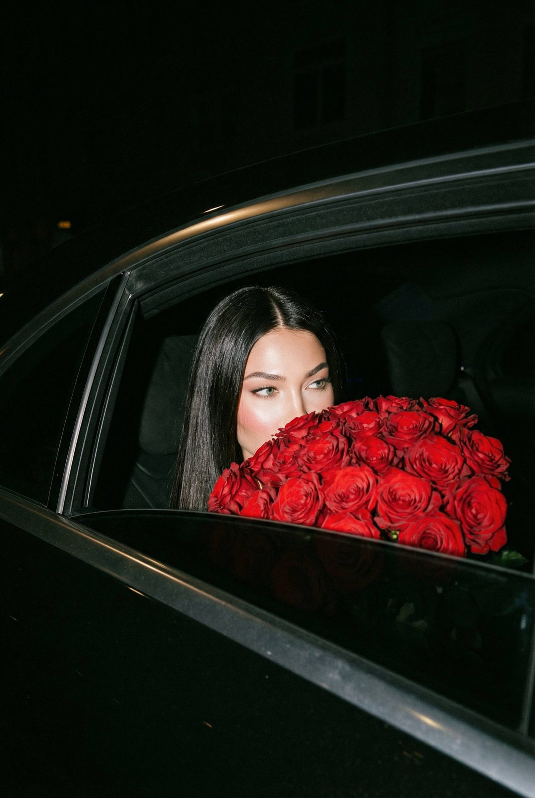 A woman in a luxury car at night, holding a large bouquet of red roses close to her face, with only her eyes visible.
