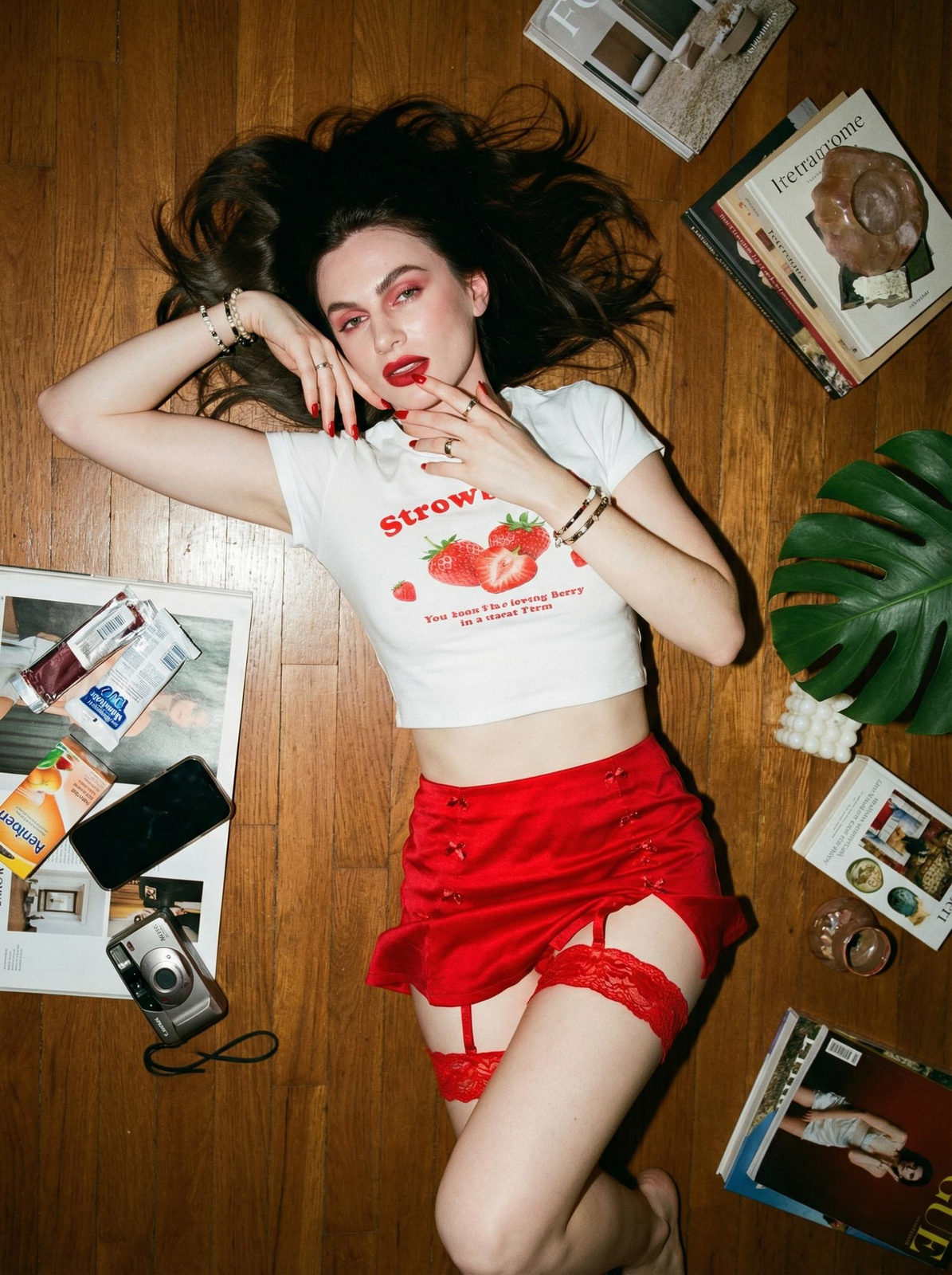 A woman lies relaxed on a brown wooden floor, wearing a strawberry crop top and red skirt, surrounded by scattered books and a green leaf, shot from above.