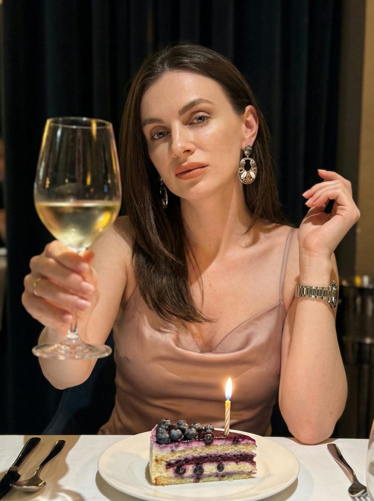 A woman in a powder-colored satin dress holding a glass of wine at a dimly lit restaurant table, with a cake and candle in front.