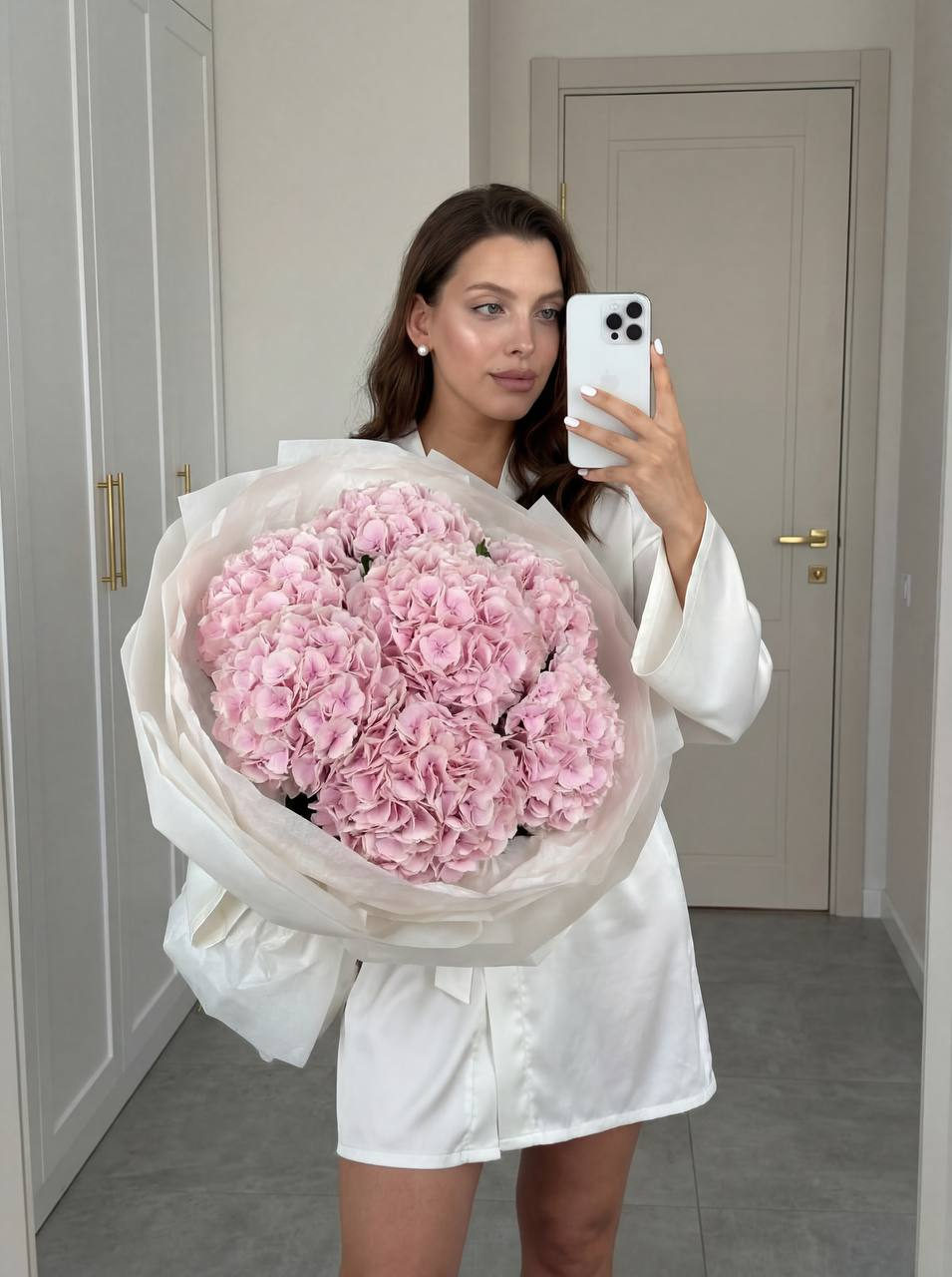 A woman in a satin robe takes a selfie in a mirror, holding a massive bouquet of soft pink hydrangeas that cover her body.