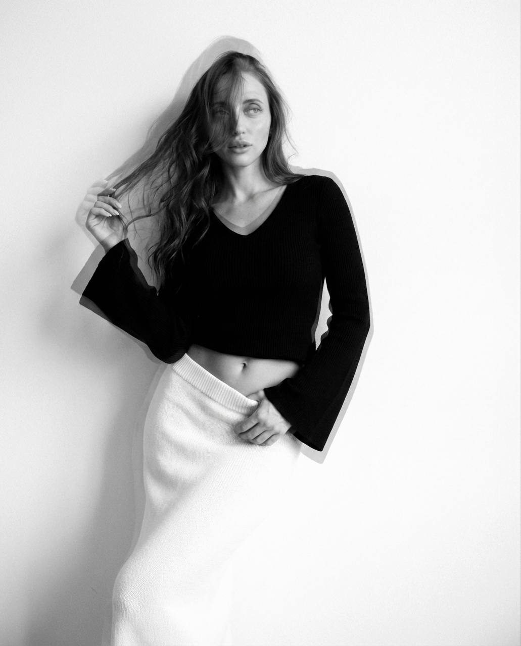 A young woman with long hair partially covering her face, wearing a black V-neck sweater and a high-waisted white textured skirt, set against a simple white wall.