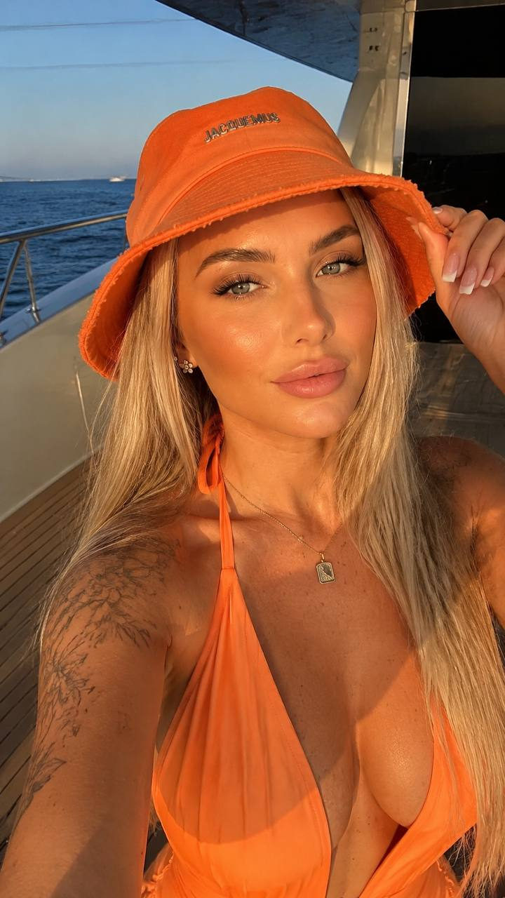A woman with a bright orange bucket hat lightly touching its brim while basking in golden hour light on a luxury yacht.