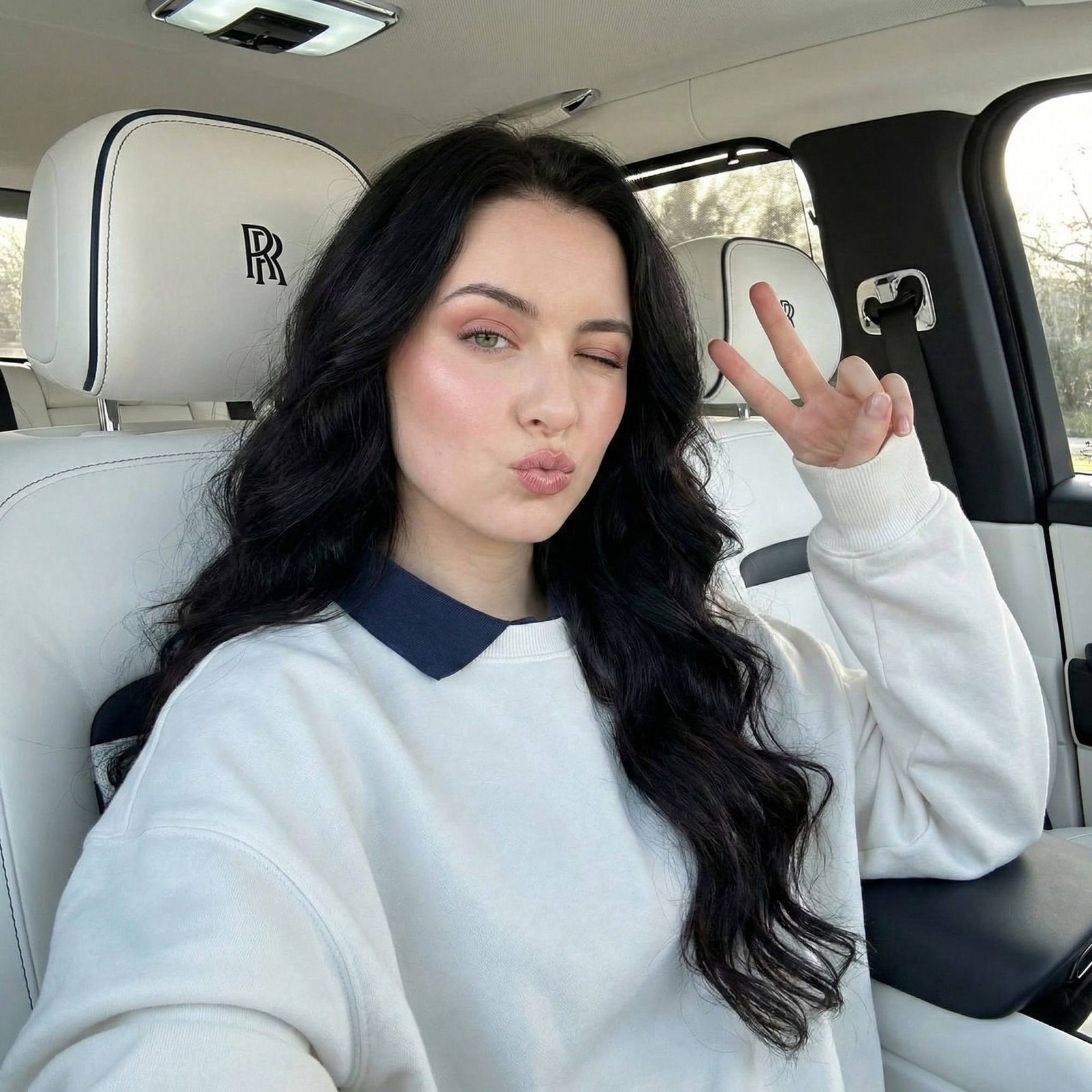 A woman with long wavy hair in a Rolls-Royce, wearing a dark blue collared sweatshirt and playfully making a peace sign.