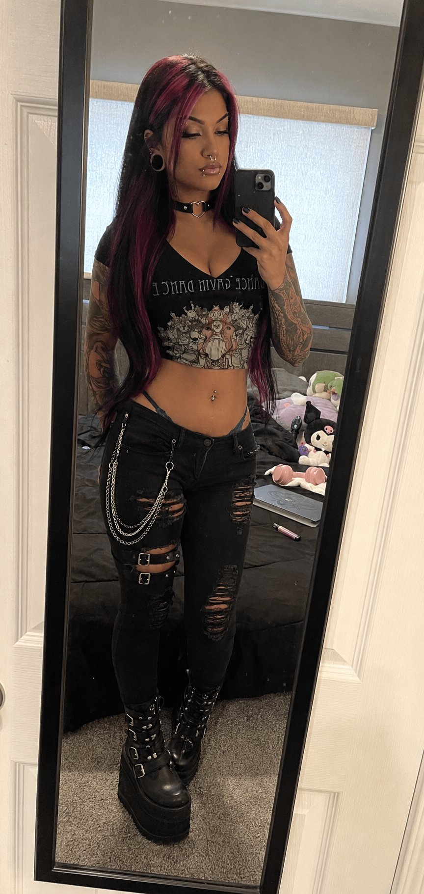 Full-length mirror selfie showing a person in a bedroom holding a smartphone with soft indoor lighting and a dark red hair look.