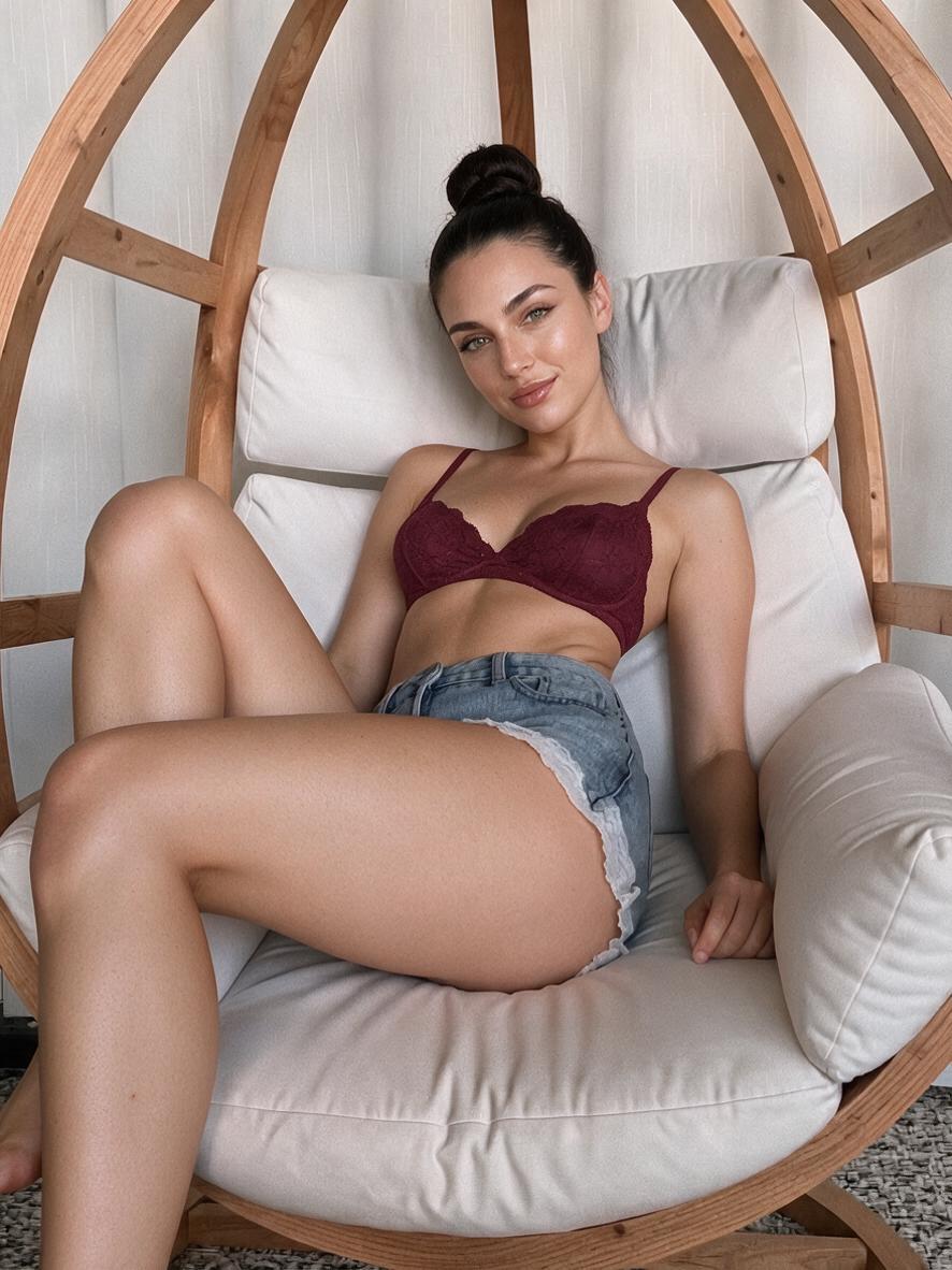 Woman in dark red lace bralette and light blue denim shorts, soft smile, seated in wooden egg chair.