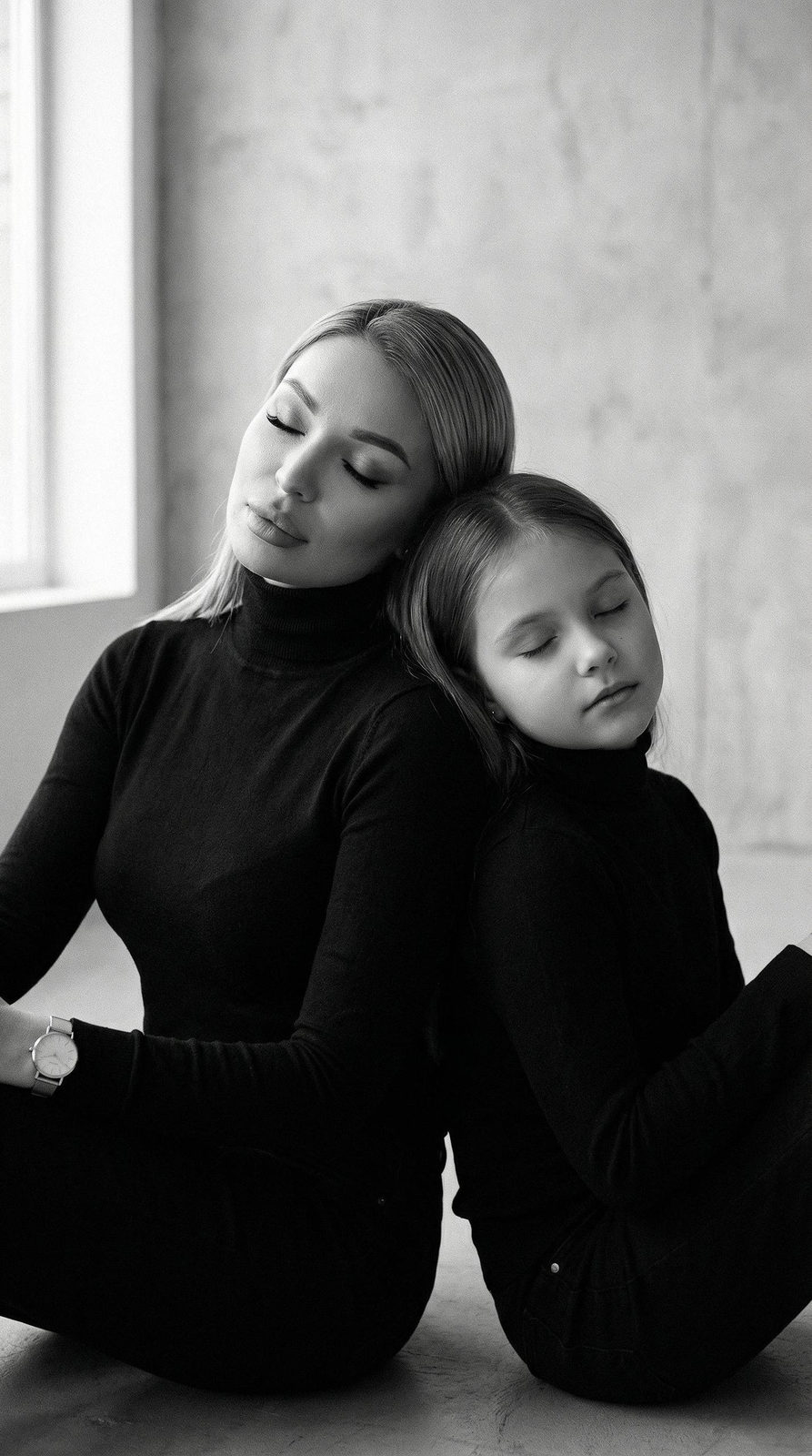 Cinematic black and white portrait of a mom and daughter sitting back to back, eyes closed, in simple black turtlenecks, soft window lighting.