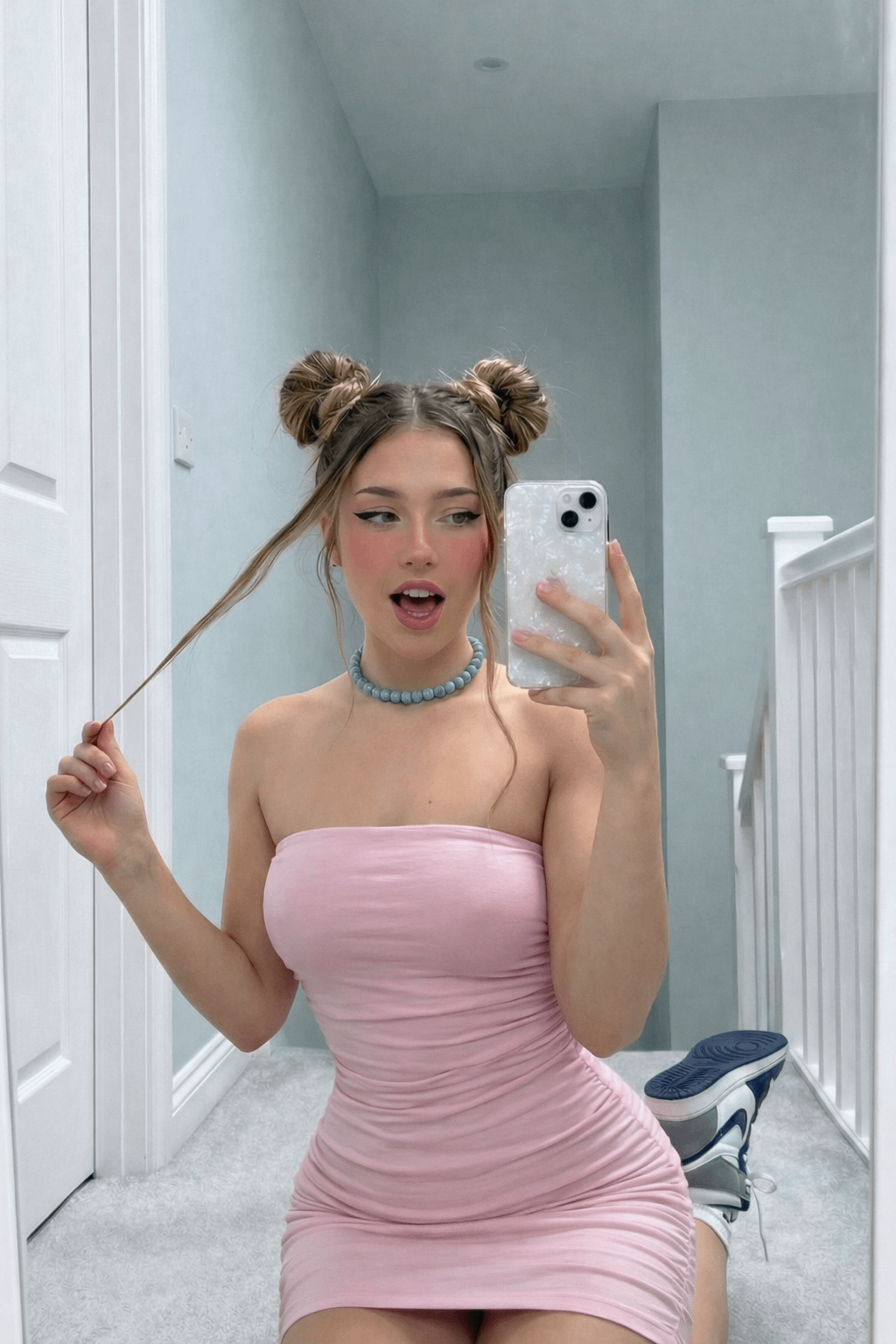 A woman in a pastel pink tube dress kneeling on a light grey carpet, taking a playful mirror selfie, with visible tongue and sharp eyeliner.