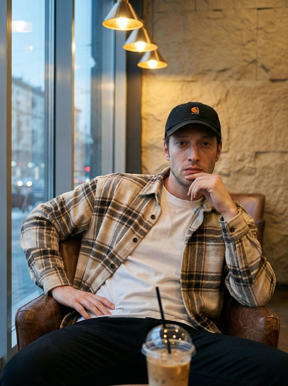 A man in a black cap and checkered shirt sits in a cozy cafe, his hand on his chin, with a warm drink beside him and a city view.