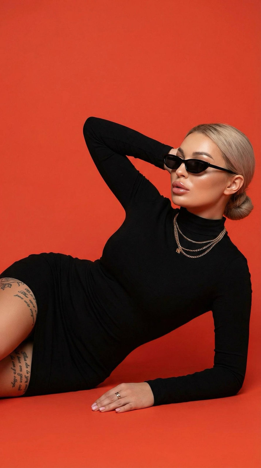 A girl in a black dress lying on her side, showcasing a chic look with gold chains and modern glasses against a bright orange background.