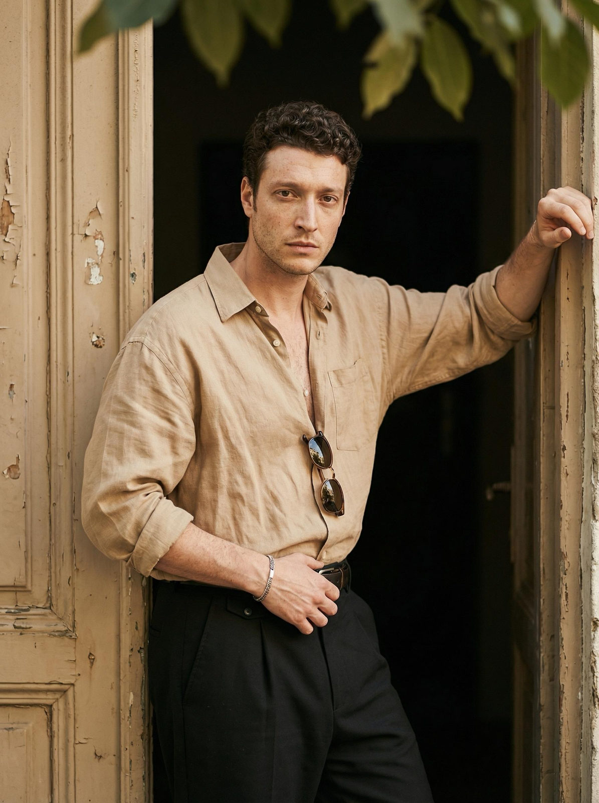 A man in a beige shirt standing in a doorway, sunglasses resting on his chest, with lush green vegetation around.