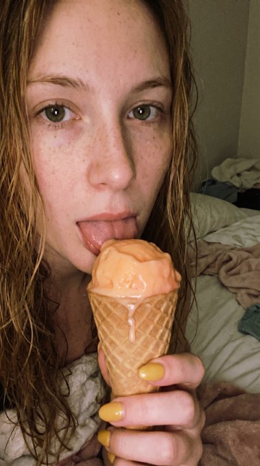 A woman holding a melting peach ice cream cone with bright yellow nails in a cozy, rumpled bed setting.