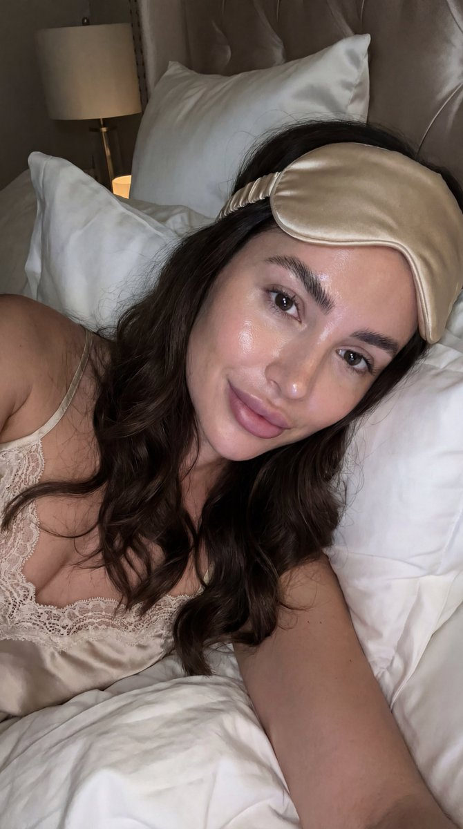 A young Hispanic woman with flawless glass skin takes a softly lit bedtime selfie, showcasing her voluminous dark waves against luxurious bedding.
