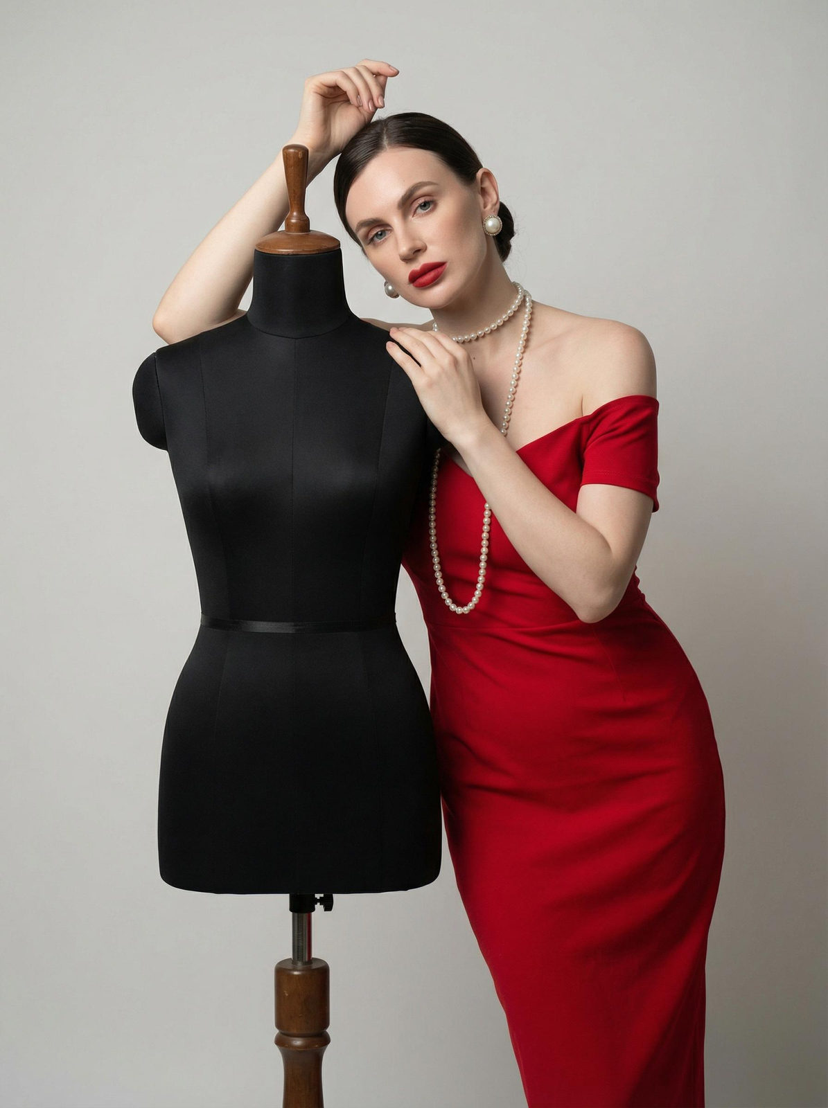 Portrait of a woman leaning on a mannequin in a red off-the-shoulder dress, styled with pearl accessories, in a minimalist studio.
