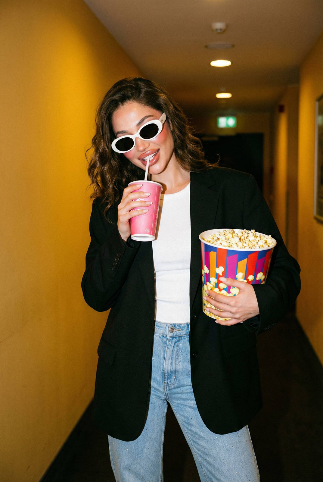A woman in an oversized black blazer holding popcorn and soda in a vibrant cinema hallway, softly lit with a stylish, casual vibe.