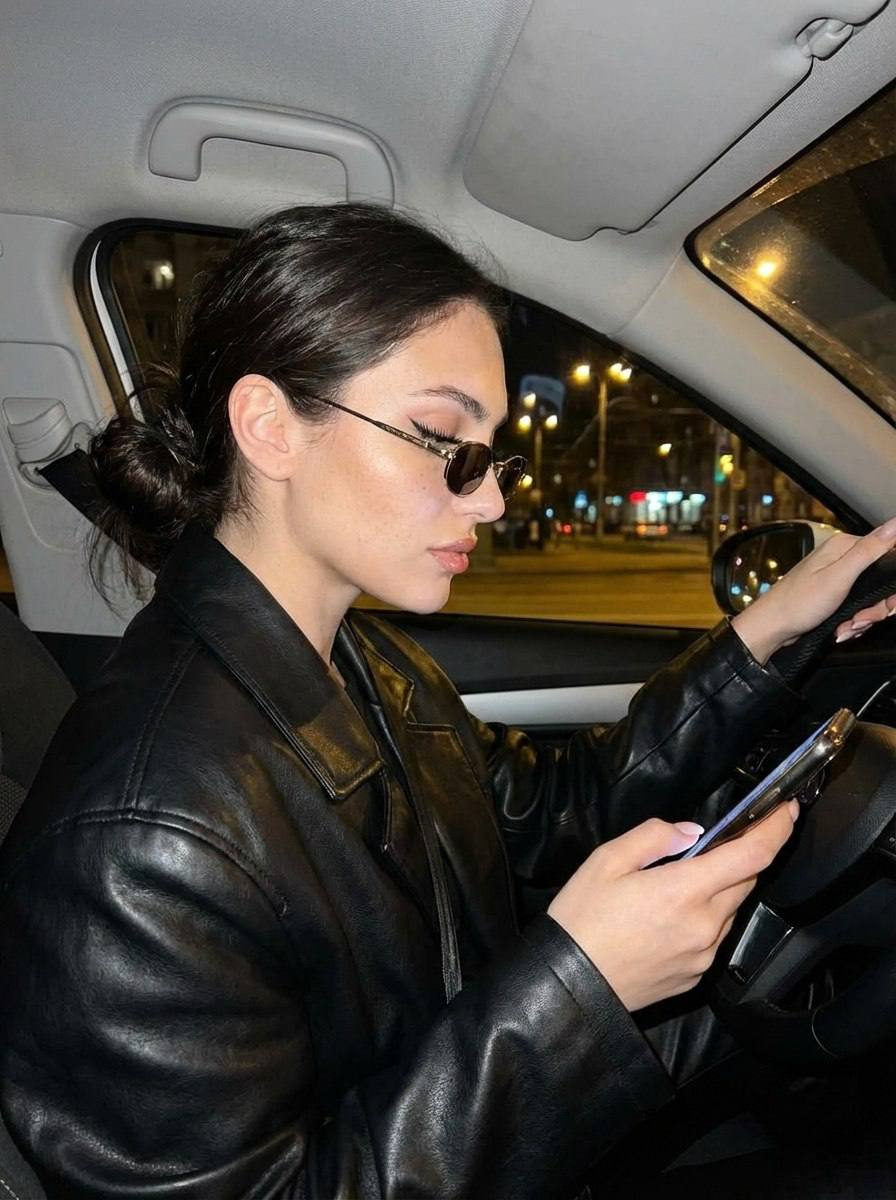 A girl in a sleek black leather jacket sitting in a car, focused on her phone, with city lights glowing outside the window.