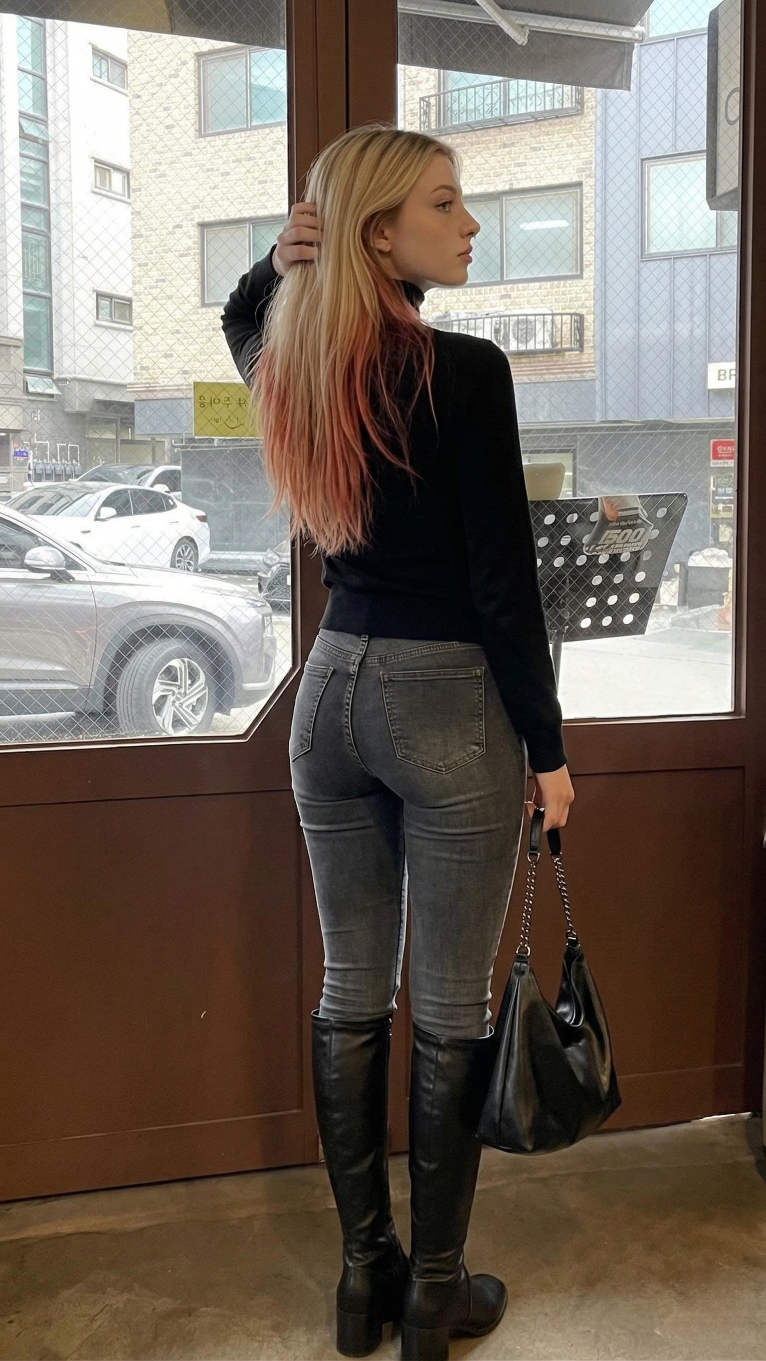 A woman in a fitted black turtleneck and denim jeans stands indoors, looking out an urban window with a city view.
