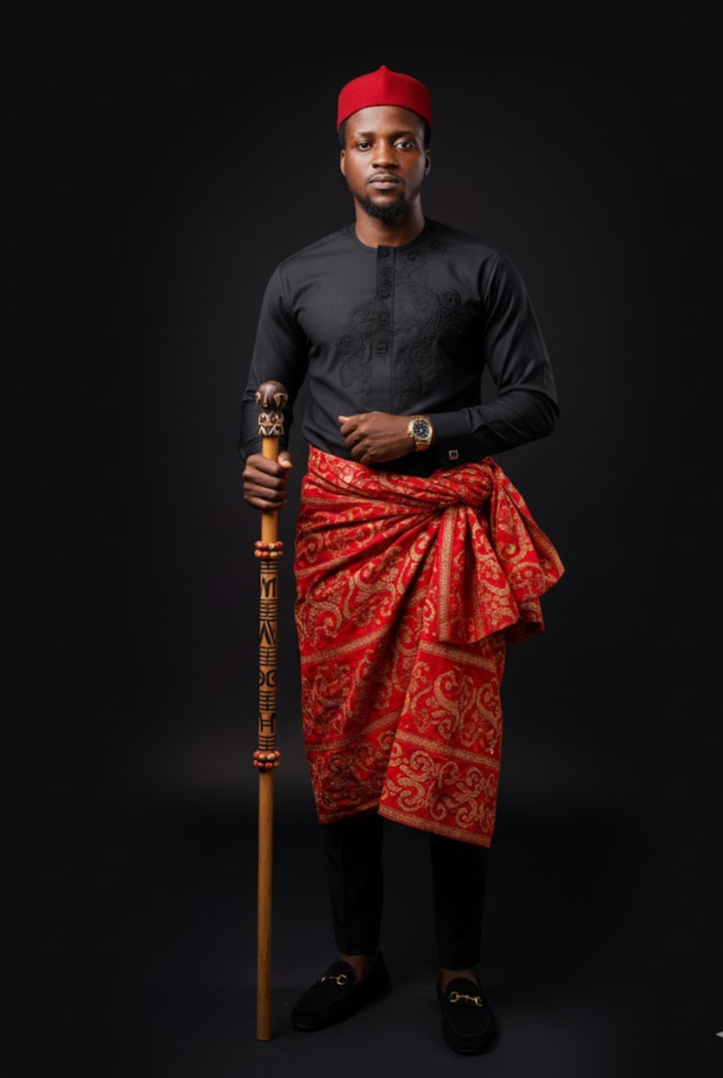 A man in a black top and red wrapper, standing confidently against a black backdrop, embodying an Igbo chieftain's elegance.