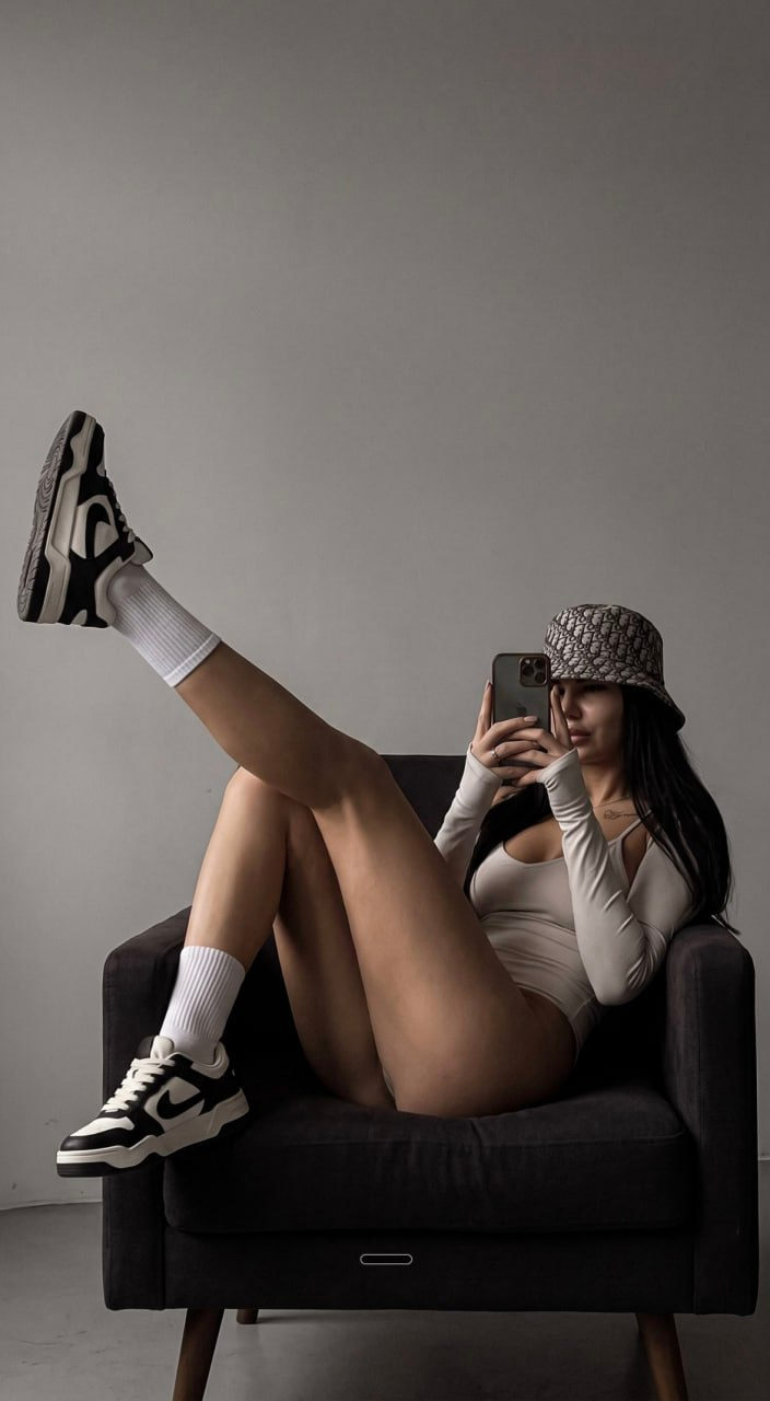 A model in a light bodysuit poses on a dark chair against a gray wall, showcasing streetwear style.