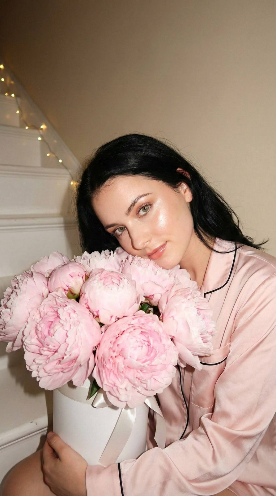 Young girl sitting on steps with cheek resting on a bouquet of soft pink peonies, wearing a light pink silky shirt
