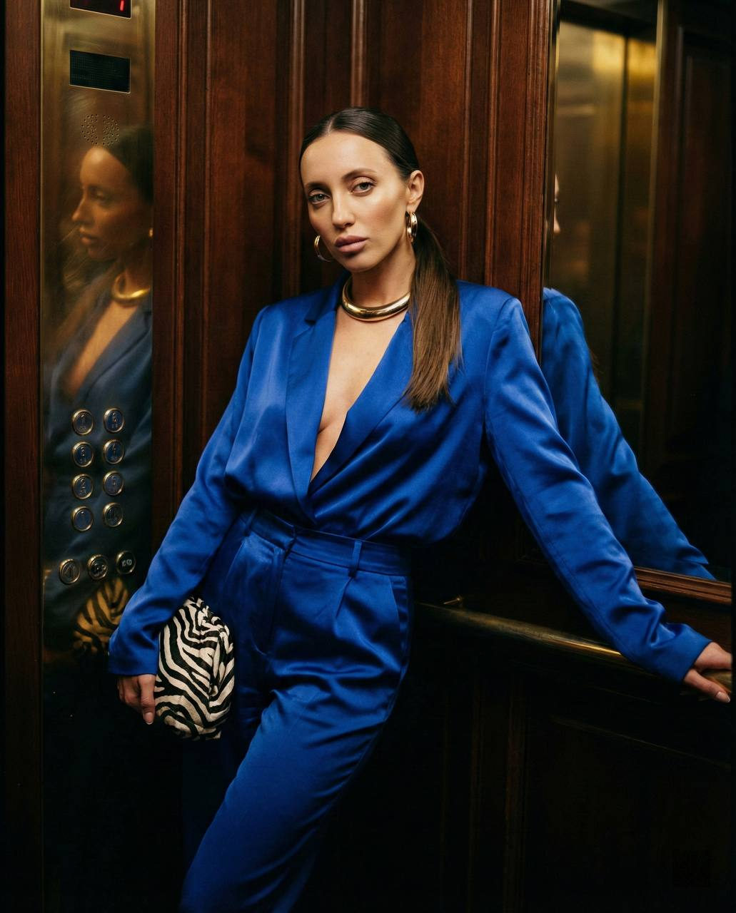 A woman in a cobalt blue satin suit poses confidently in a vintage elevator, dramatic lighting highlighting her silhouette.