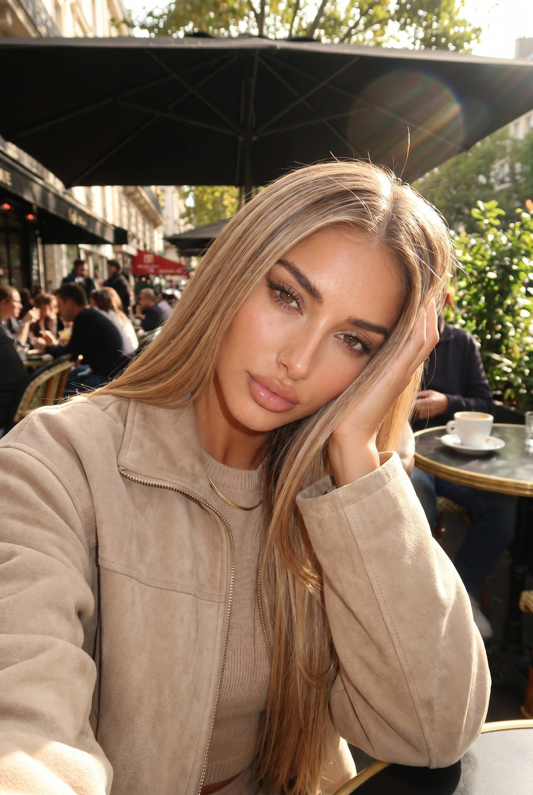 A woman with long sleek hair resting her cheek on her hand while gazing at the camera, seated at an outdoor café table.